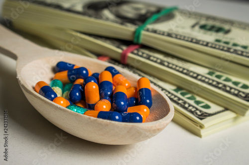 Many colored capsules in a wooden spoon on a white table with packs of dollars