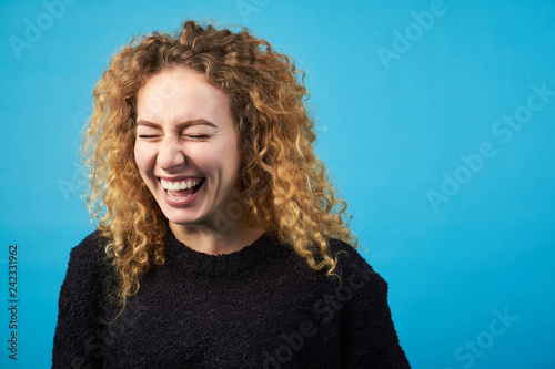 Funny attractive redhead curly girl laughing  with closed eyes on blue background.