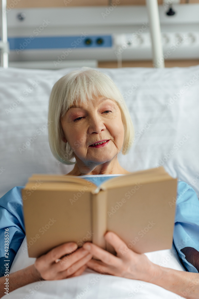 selective focus of senior woman with grey hair lying in bed and reading book in hospital selective focus of senior woman with grey hair lying in bed and reading book in hospital