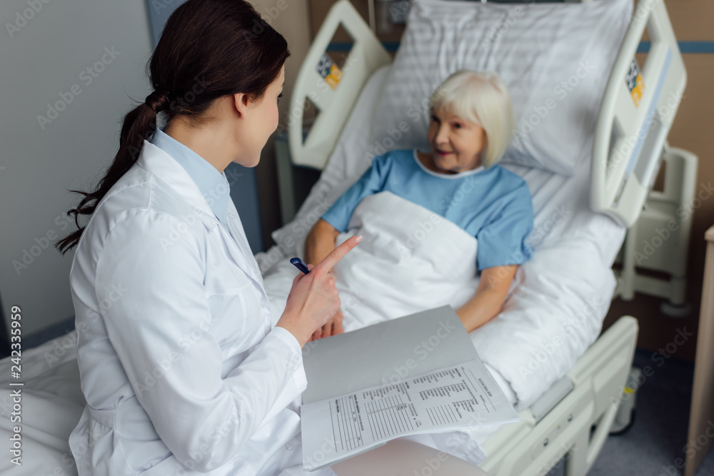 Fototapeta premium female doctor sitting on bed, holding diagnosis and pointing with finger while senior woman lying in bed in hospital