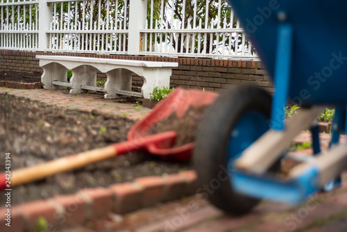 Spring time in the garden with white fence and bench with soft focus on fore ground with brick lined garden bed, a red shovel and blue wheelbarrow.  