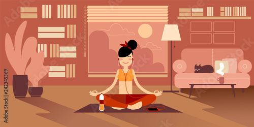 A Yoga girl in headphones meditates at home, sitting on a pilates mat in an easy sukhasana pose. Nearby there is a bottle of water, a cup of tea and a cat on the sofa. photo
