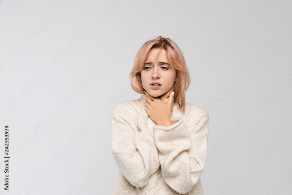 Horizontal studio portrait of young unhealthy blonde woman in warm sweater holding her throat/suffers from cold and flu, severe sore throat, feeling sick, chest pain, medicine and healthcare concept.
