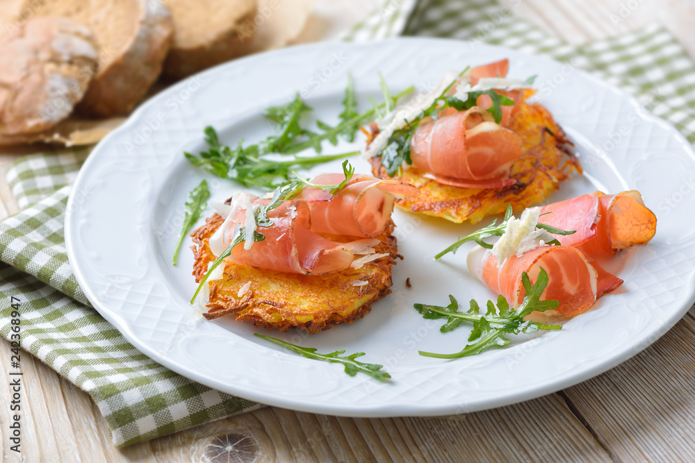 Deftige Kartoffelrösti mit Südtiroler Speck, Parmesan und Rucola rustikal serviert – Potato rosti with South Tyrolean bacon, parmesan cheese and rocket salad served on a white plate