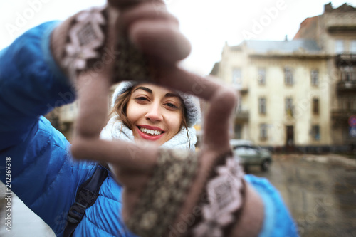 portrait of a young smiling attractive woman in warm winter clothes at cloudy day on the street background. woman shows a frame from hands like photo. Photo Frame Hands Made By A Hipster young girl