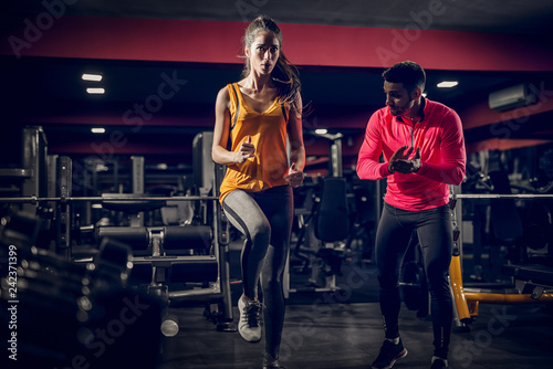 Fit beautiful focused girl warming up in a gym with her personal trainer by her side.