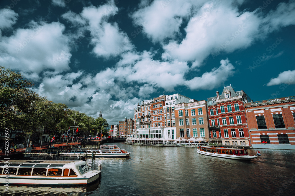 Classical Amsterdam cityscape. Cruise boats floating on the channel, river side promenade, cafes, typical Dutch architecture. Urban scene and white fluffy clouds