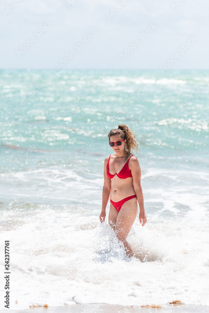 Young girl woman in red wet swimsuit bikini on beach during sunny day in Miami, Florida standing with water shore wave crashing Young girl woman in red wet swimsuit bikini on beach during sunny day in Miami, Florida standing with water shore wave crashing