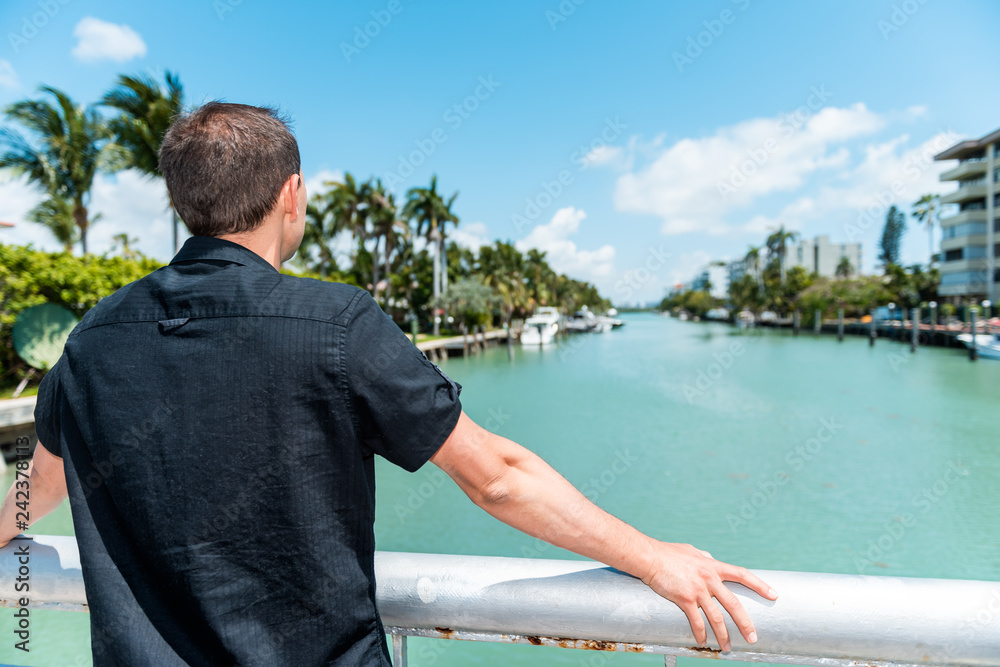 Young man back wearing black shirt standing leaning on bridge railing in Bal Harbour, Miami Florida with green ocean Biscayne Bay