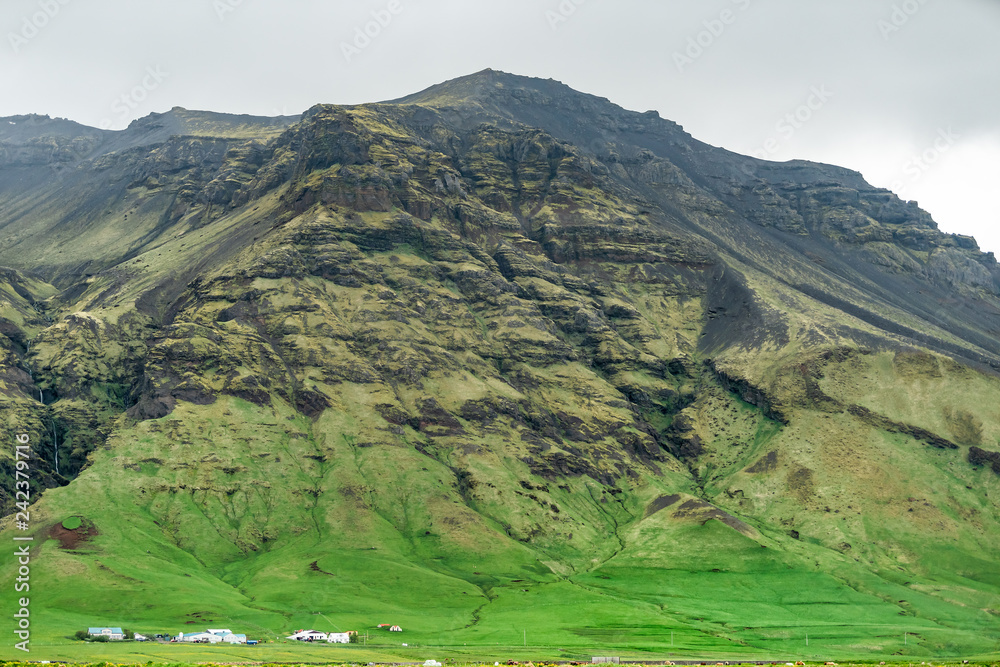 Snowcapped landscape view of green mountain on cloudy day, southern ring road or golden circle and farm houses near Skogafoss Skogar waterfall