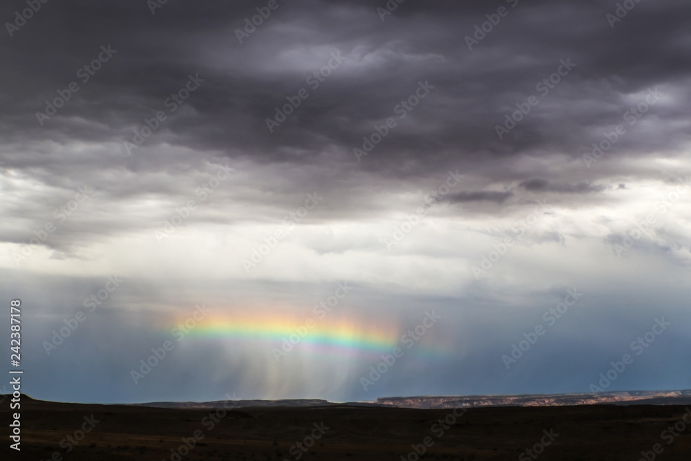 Horizontal rainbow across the middle of a rain squall near the horizon in desert with distant cliffs lit up and foreground dark - very dramatic stormy sky near Salt Lake USA