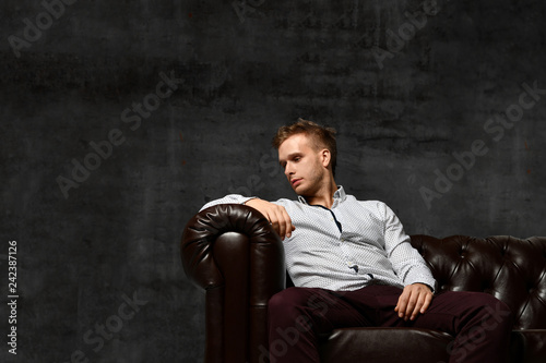 Young business man sitting in leather Chester armchair sofa in casual shirt thinking