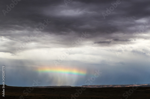 Horizontal rainbow across the middle of a rain squall near the horizon in desert with distant cliffs lit up and foreground dark - very dramatic stormy sky near Salt Lake USA