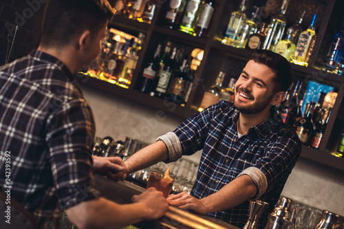 Young bartender standing at bar counter with customer talking happy