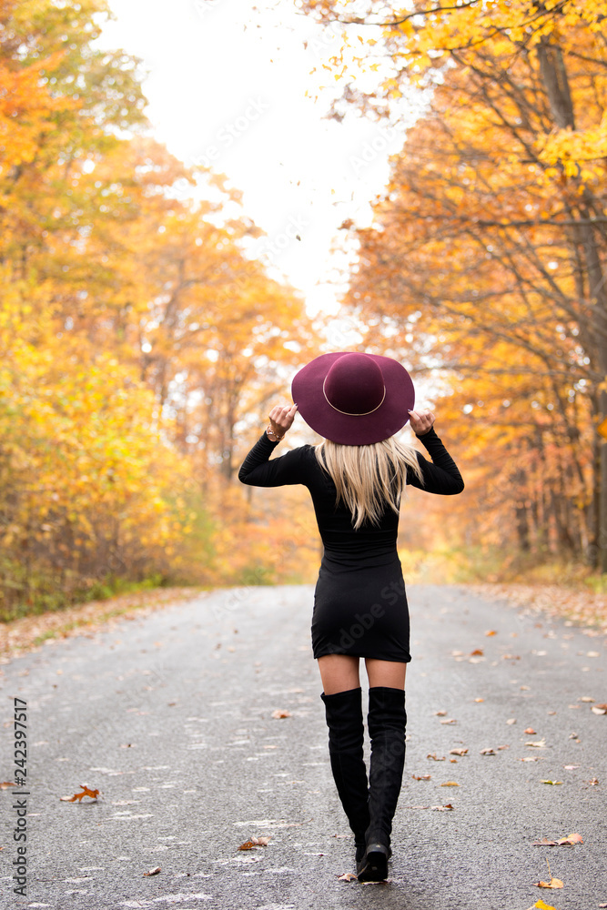 Sexy young blonde woman in stunning black dress and over the knee boots  wears a colorful hat standing on deserted country woods in fall - autumn  fashion Photos | Adobe Stock
