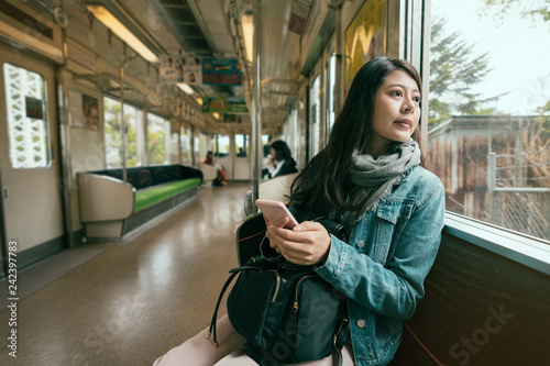 asian travel woman inside subway train looking out beautiful view from window. young girl holding smart phone sitting on seat in metro. fashion chinese woman commute in tour trip wear scarf spring