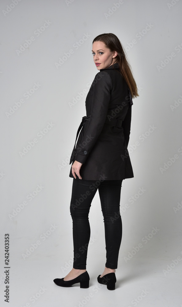  full length portrait of a brunette girl wearing long black coat, standing pose with back to the camera, on grey studio background.