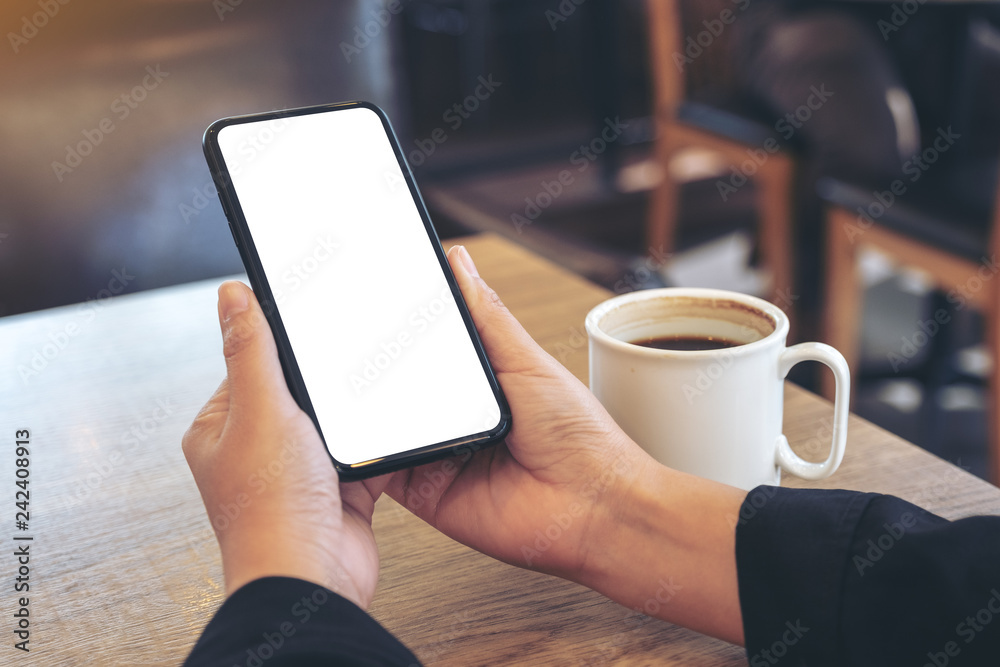 Mockup image of hands holding white mobile phone with blank screen with coffee cup on wooden table in cafe