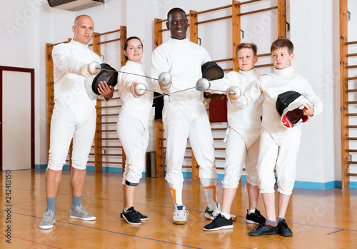 Group portrait of young fencers with coaches holding rapiers in training room