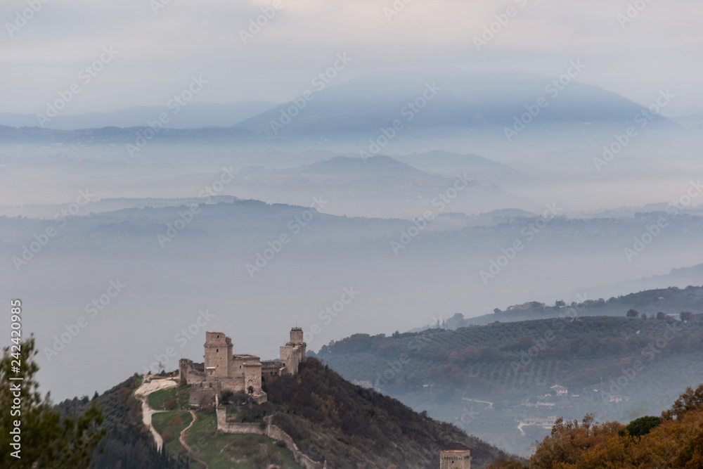 Beautiful view of Rocca Maggior castle (Assisi, Umbria, Italy) over a sea of fog with distant hills in the background and orange and green trees in the foreground