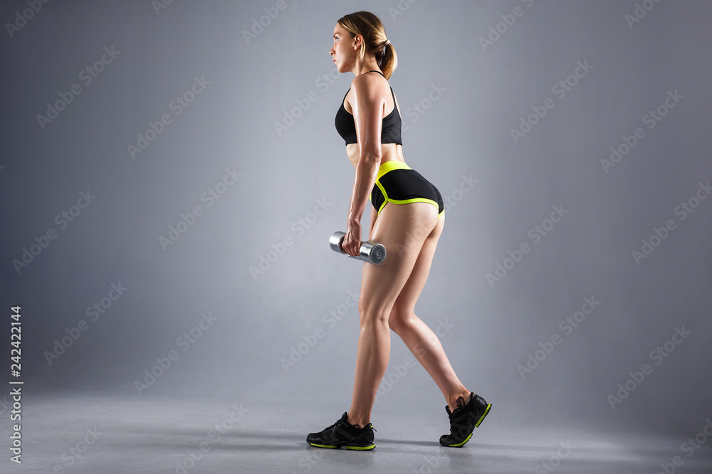 Beautiful young woman powerlifters in a sport suit and sneakers doing slopes with dumbbells on a gray background