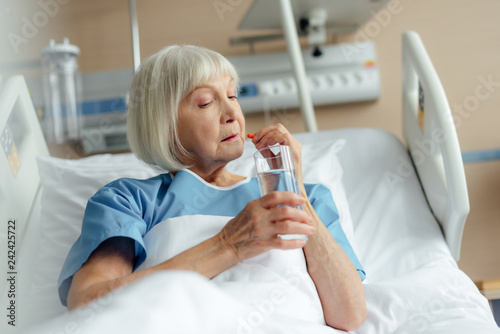 selective focus of senior woman lying in bed, holding glass of water and taking pill in hospital