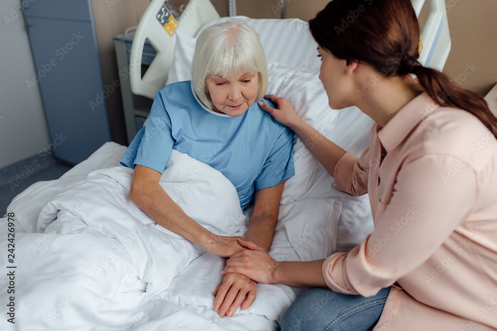 daughter sitting near senior mother on bed and holding hands in hospital daughter sitting near senior mother on bed and holding hands in hospital