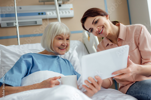 happy senior woman and daughter using digital tablet in hospital bed