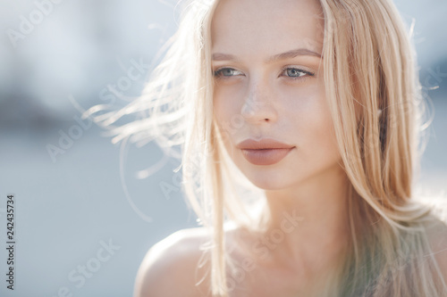 A young beautiful woman with long, straight hair and gray eyes, beautiful makeup, pink plump lips, sweet smile, blonde, spending her time outdoors in the summer in the city, posing in the sunlight