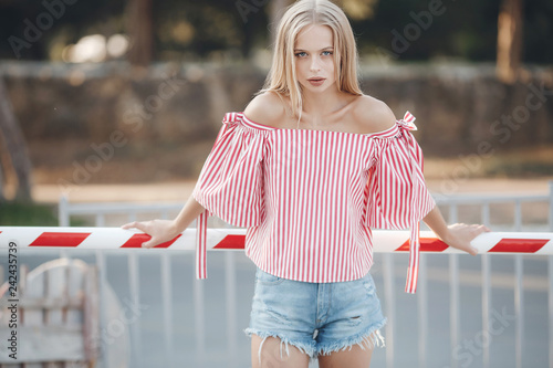 A young beautiful woman with long, straight hair and gray eyes, beautiful makeup, pink plump lips, sweet smile, blonde, spending her time outdoors in the summer in the city, posing in the sunlight