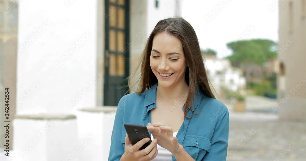 Happy woman walking towards camera and browsing cellphone in the street Happy woman walking towards camera and browsing cellphone in the street