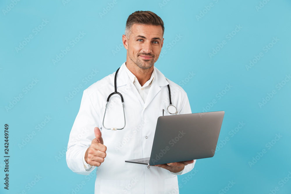 Photo of mature man wearing white medical coat and stethoscope holding laptop, standing isolated over blue background Photo of mature man wearing white medical coat and stethoscope holding laptop, standing isolated over blue background