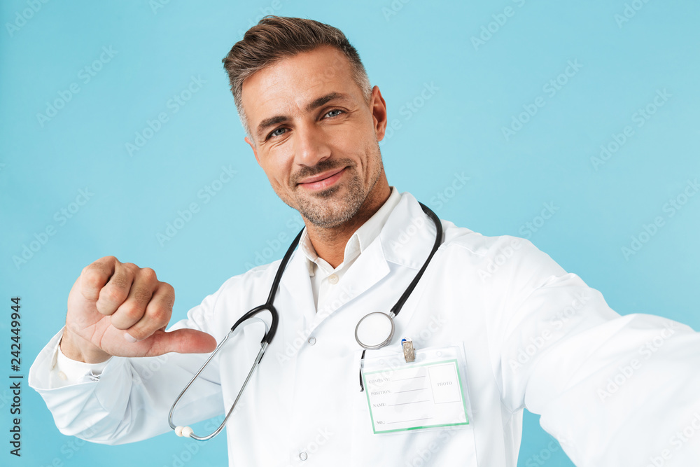 Portrait of caucasian medical doctor with stethoscope taking selfie photo, while standing isolated over blue background Portrait of caucasian medical doctor with stethoscope taking selfie photo, while standing isolated over blue background