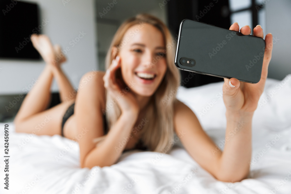 Image of pretty woman 20s wearing luxury lingerie smiling, while lying in bed on white linen in cozy flat and using smartphone Image of pretty woman 20s wearing luxury lingerie smiling, while lying in bed on white linen in cozy flat and using smartphone