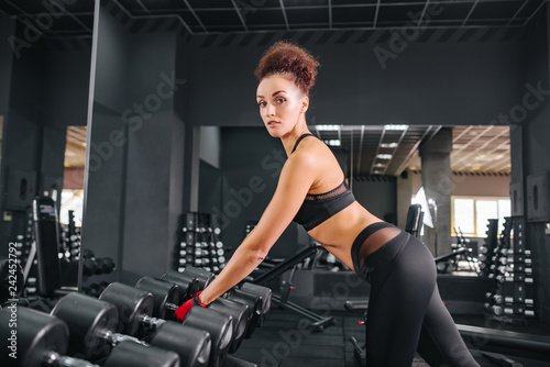 Attractive fitness woman in black sportswear top and leggins with perfect body posing in the gym. Sport workout exercises with dumbbels. Still dumbells.