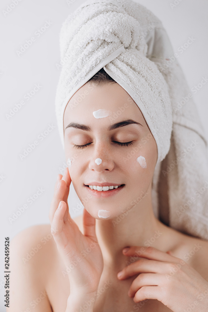 Young happy woman wrapped with bath towel applying cream to her face for skin care. Skin care concept. Skin care and beauty concept. Place for text Young happy woman wrapped with bath towel applying cream to her face for skin care. Skin care concept. Skin care and beauty concept. Place for text