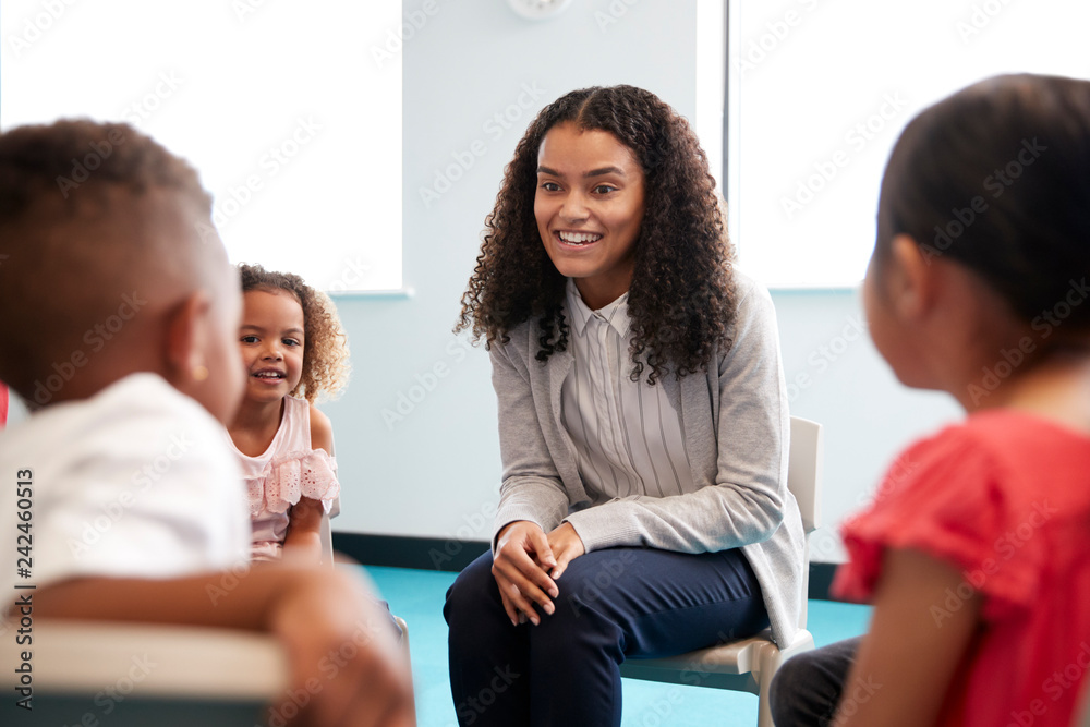 Infant school children sitting on chairs in a circle in a classroom talking to their smiling female teacher, close up, selective focus Infant school children sitting on chairs in a circle in a classroom talking to their smiling female teacher, close up, selective focus