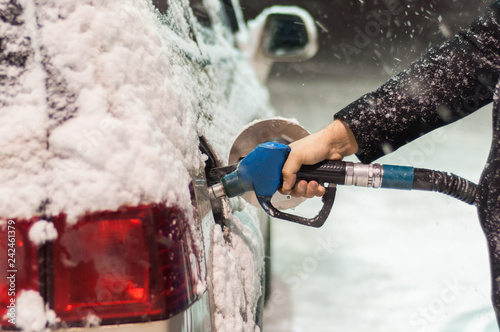 A man in a black coat fills his car at a gas station on a snowy winter evening. Hand holding a blue refueling gun..