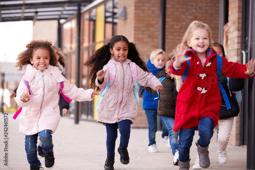 Three happy young school girls wearing coats and carrying schoolbags running in a walkway with their classmates outside their infant school building, close up