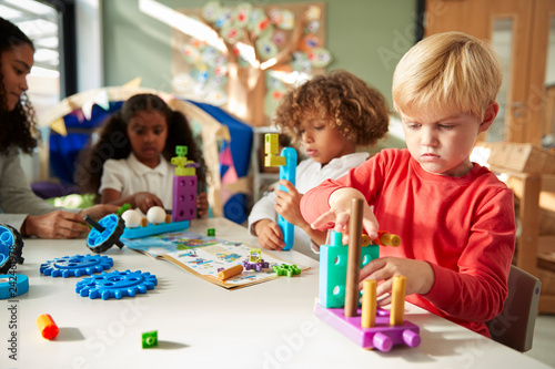 Infant school boy sitting at a table using educational construction toys with his classmates, close up