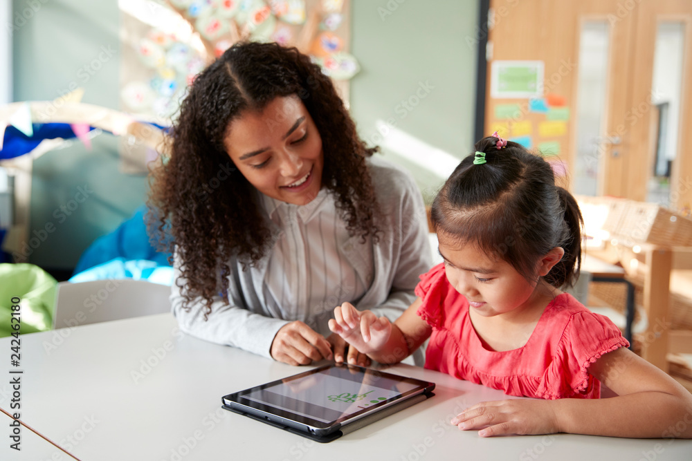 Female infant school teacher working one on one with a Chinese schoolgirl, sitting at a table in a classroom using a tablet computer, close up Female infant school teacher working one on one with a Chinese schoolgirl, sitting at a table in a classroom using a tablet computer, close up