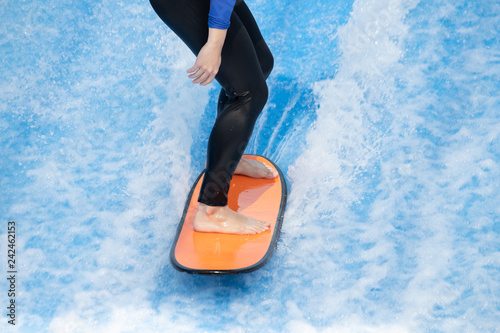 Photography woman surfing in beach wave simulator attraction of water park, wearing black swiming suit balancing on orange surfboard in fake wave