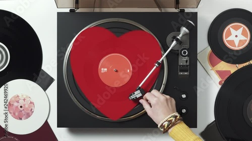 A red heart vinyl record turntable on a white table with plates. Included gramophone, torque. The hand of a girl DJ with bright accessory puts a stylus with a needle on a vinyl record. Valentine's Day photo