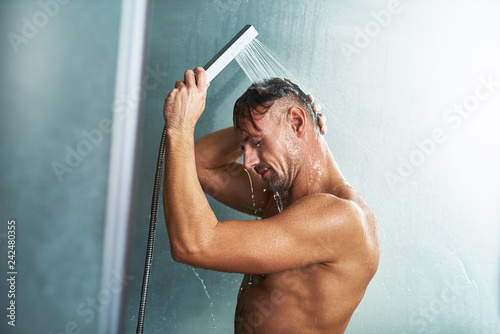 Handsome young man using handheld shower head while washing himself