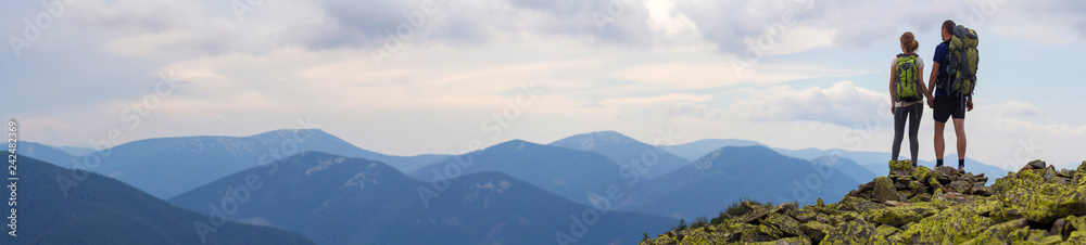 Back view of young tourist couple with backpacks, athletic man and slim girl stand holding hands on rocky mountain top enjoying mountain panorama. Tourism, traveling and healthy lifestyle concept.