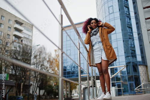 Attractive curly african american woman in brown coat posed near railings against modern multistory building speaking on mobile phone.