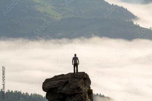 Silhouette of athletic climber tourist on high rocky formation on mountain valley filled with white puffy clouds and fog and covered with evergreen forest mountain slopes under clear sky background.