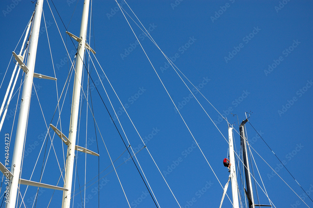 Masts of sailing boats, with a spreader, shrouds. A sailor is climbing up on a mast. A blue sky in the background. Masts of sailing boats, with a spreader, shrouds. A sailor is climbing up on a mast. A blue sky in the background.