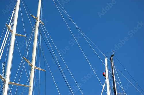 Masts of sailing boats, with a spreader, shrouds. A sailor is climbing up on a mast. A blue sky in the background.