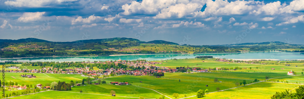A beautiful landscape panorama photo of the village Schwangau with its green fields, the turquoise blue Forggensee lake and the natural surrounding in the district of Ostallgäu in Bavaria, Germany.